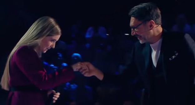 Sergey Shnurov took Ragda Khanieva by the hand in "The Voice" song show. Screenshot of the video on YouTube Голос / The Voice Russia https://www.youtube.com/watch?v=B5BVyY6glCQ&amp;feature=youtu.be